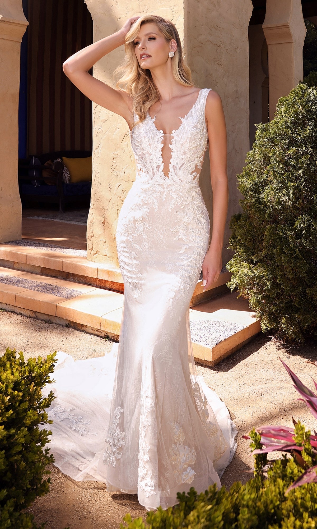 A woman wearing the Ladivine A1072W Long Off-White Lace Bridal Gown, featuring a fitted sleeveless design with a sheer bodice and flowing train, stands outdoors by stone columns and lush greenery.